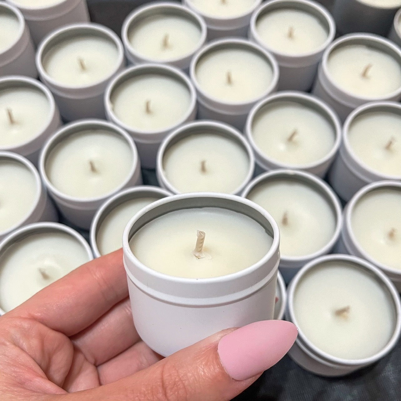 Hand holding a white tin soy candle with no label, surrounded by matching candles—perfect for minimalist or custom-branded designs.