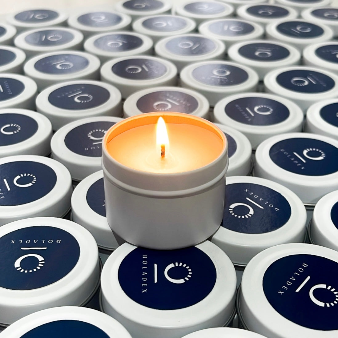 Custom branded candle tins for Roladex, with sleek navy labels and a glowing flame adding warmth and ambiance.