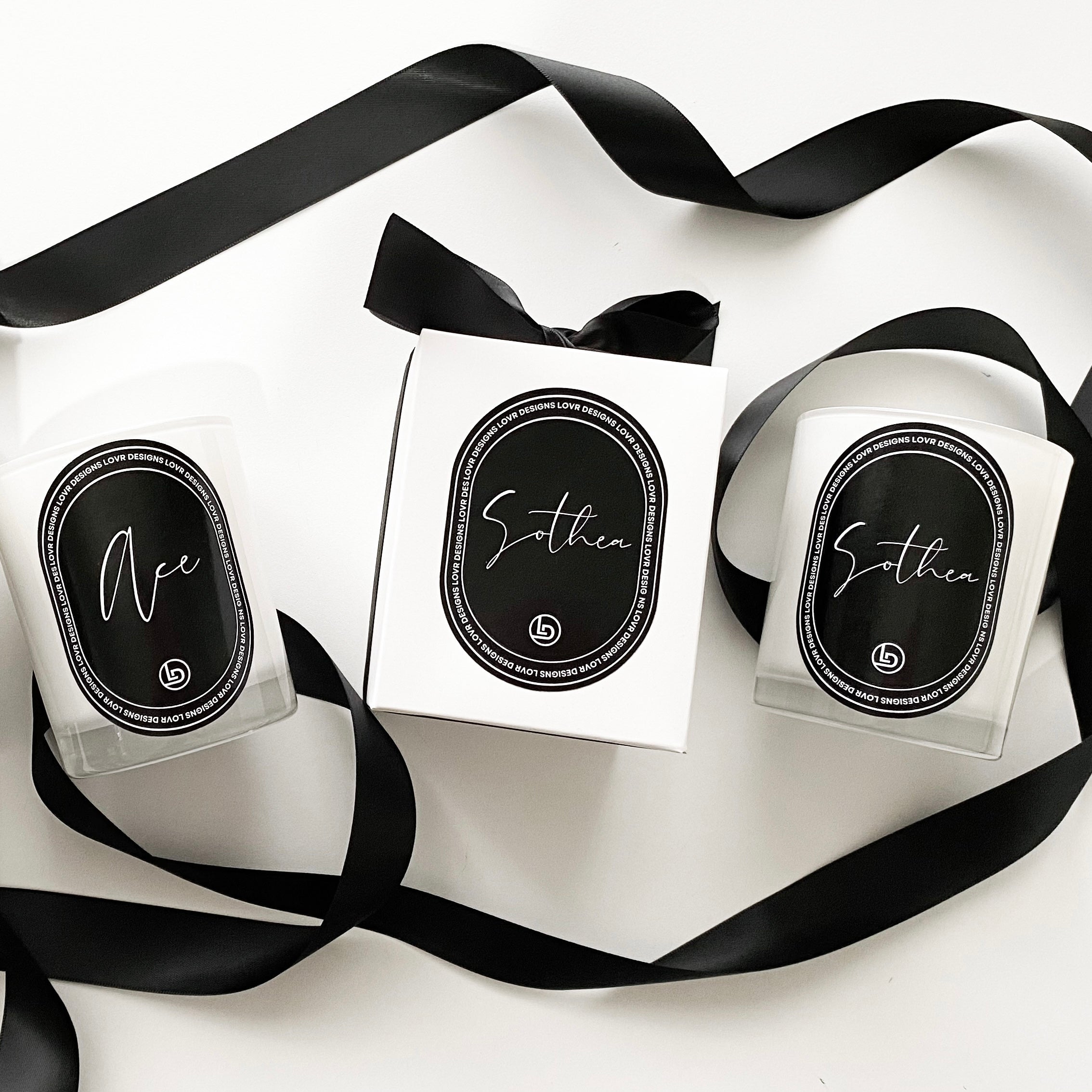 Flat-lay of Lovr Designs personalised scented soy candles labeled ‘Ace’ and ‘Sothea’ Nmes alongside a matching gift box and flowing black ribbon. Hand-poured in Melbourne, these monogrammed candles make a luxurious custom gift.