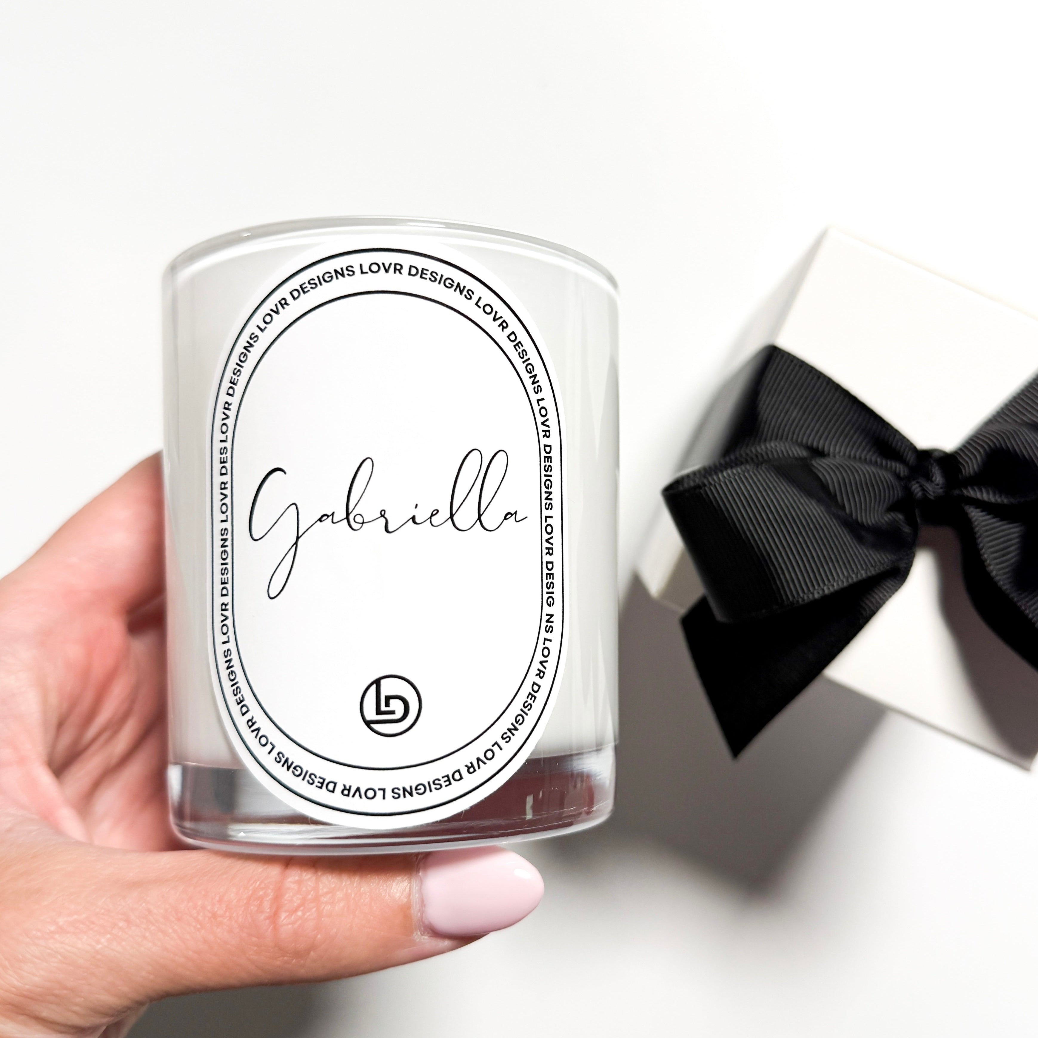 Close-up of a hand holding a Lovr Designs personalised ‘Gabriella’ Name candle jar, featuring a sleek white label with cursive lettering. Premium soy wax candle made in Melbourne, ready to gift in its matching box.