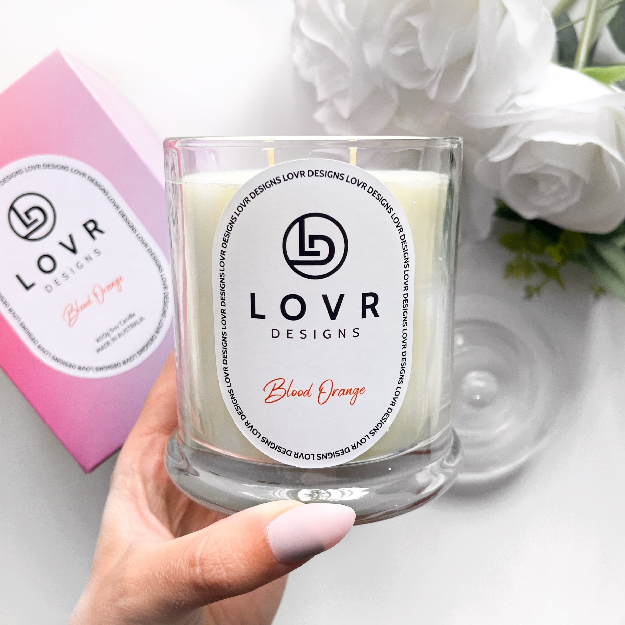 Hand holding Lovr Designs Blood Orange soy candle with gift box and white roses in background.