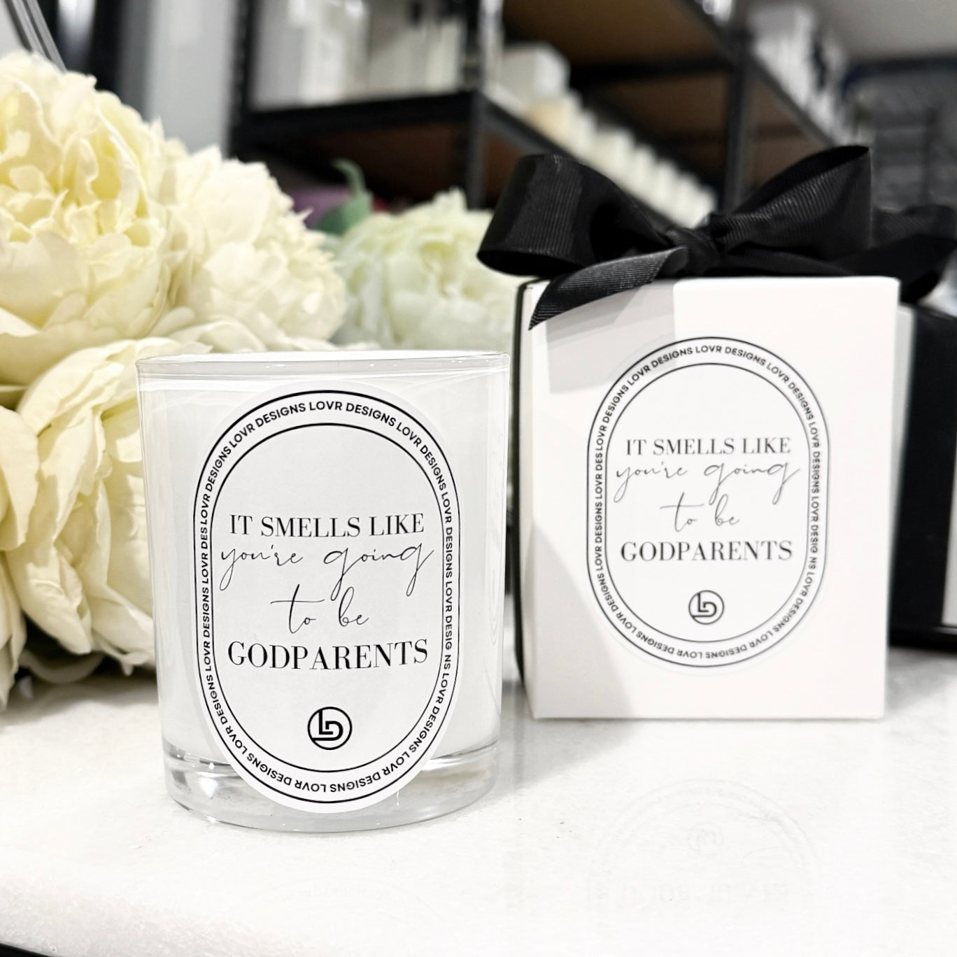 Candle and packaging with text 'It smells like you're going to be godparents' on a white surface with flowers in the background.